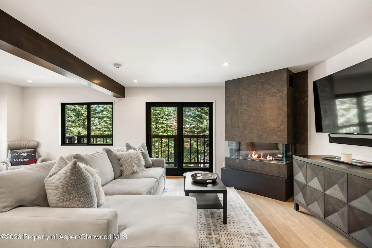 Living room with gray sectional sofa, fireplace, TV, and balcony doors with a view of trees.