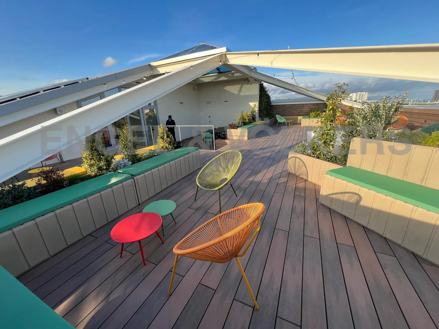 Rooftop patio with wooden deck, colorful chairs, and built-in benches with green cushions. Modern architecture and city views in the background.