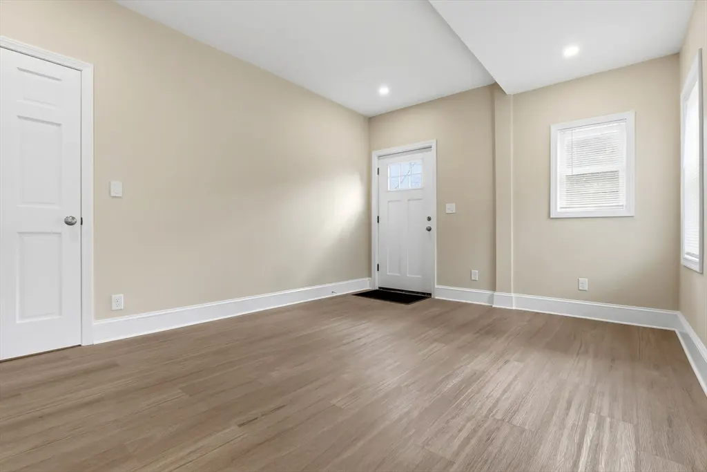 An empty room with light beige walls, light wood floors, and white trim. A white door and window are visible.