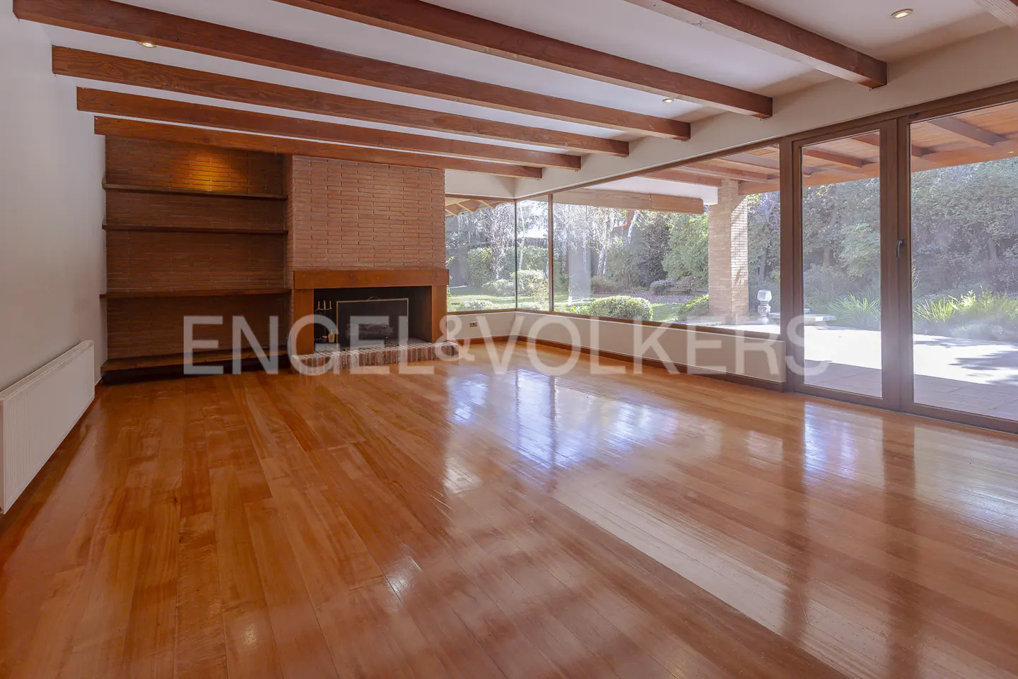 Empty living room with wood floors, exposed beams, brick fireplace, and large windows overlooking a green yard.