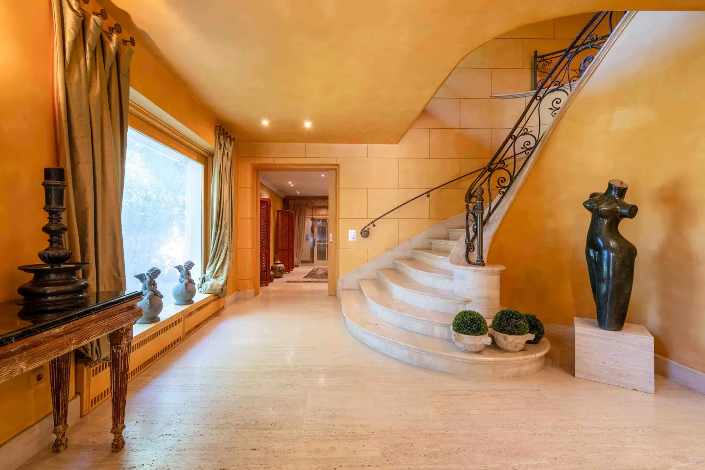 Elegant foyer with a curved stone staircase, wrought iron railing, warm yellow walls, and a modern abstract sculpture.