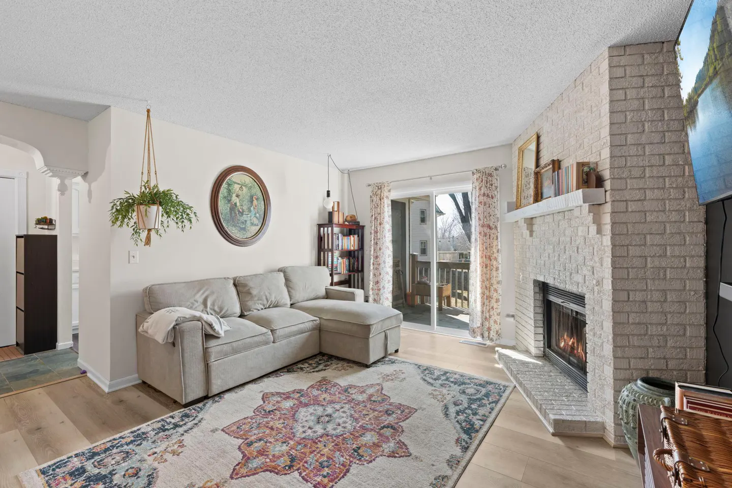Living room with a gray sectional sofa, patterned rug, and white brick fireplace with a burning fire. Sliding glass doors lead to a balcony.