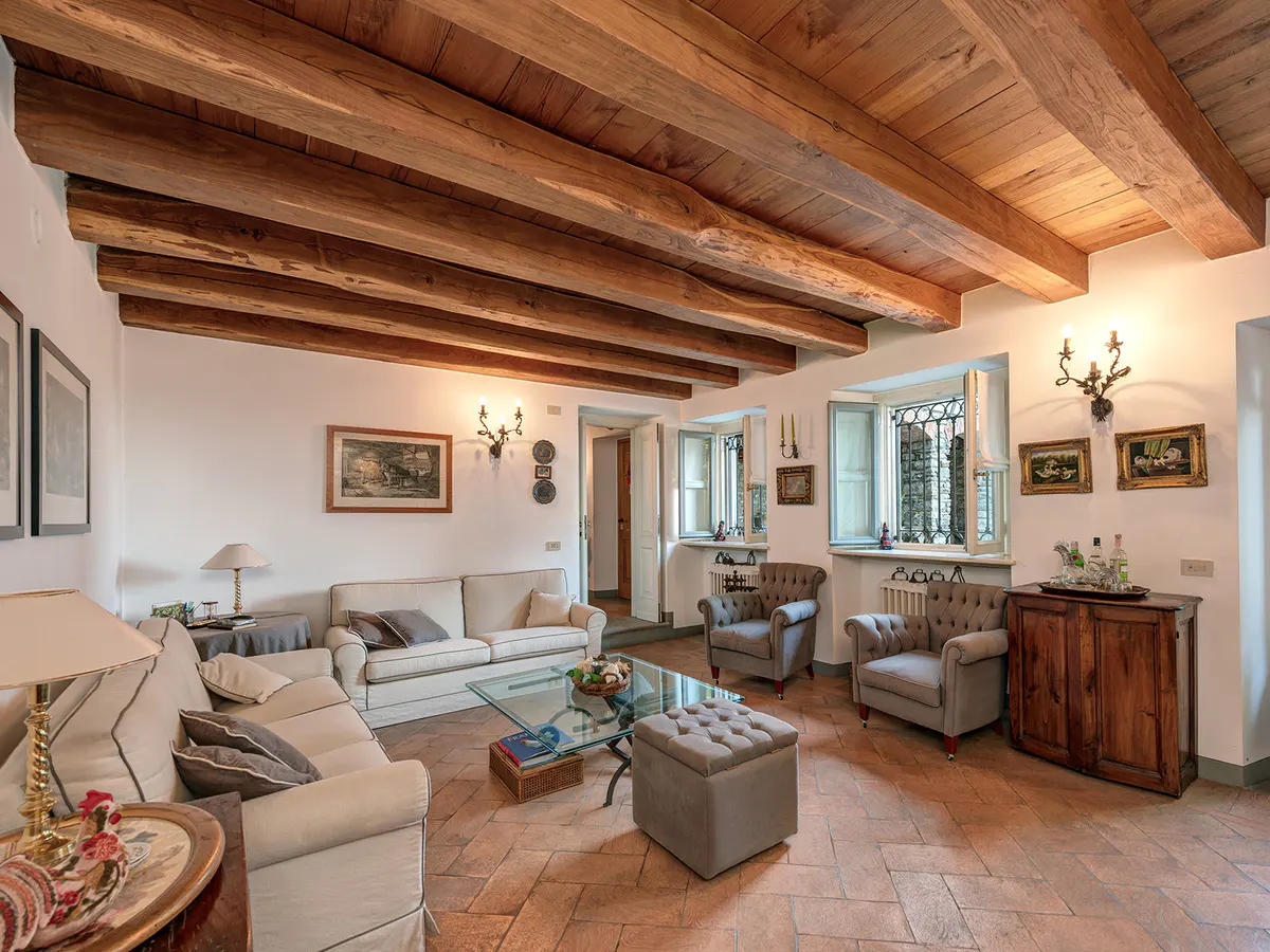 Living room with exposed wood beams, white walls, and terracotta tile floor. Sofas, chairs, glass table, and wood cabinet furnish the space.
