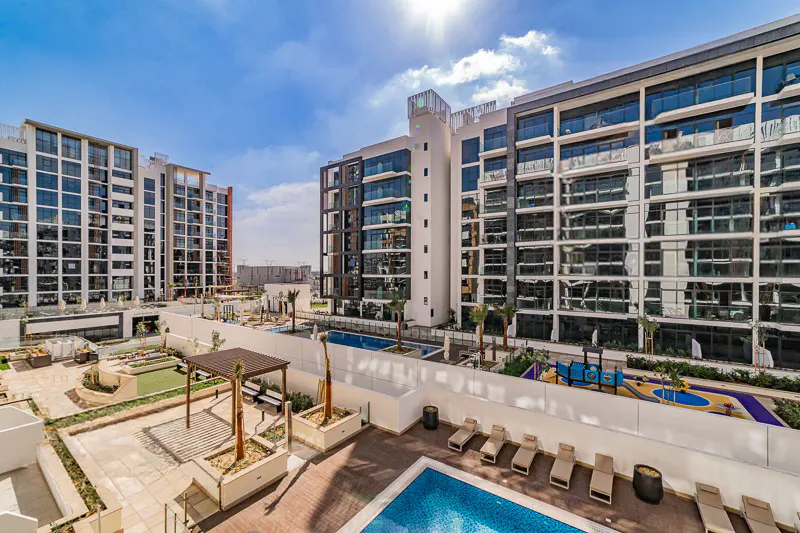Modern apartment complex with a swimming pool, lounge chairs, and a playground under a bright blue sky.