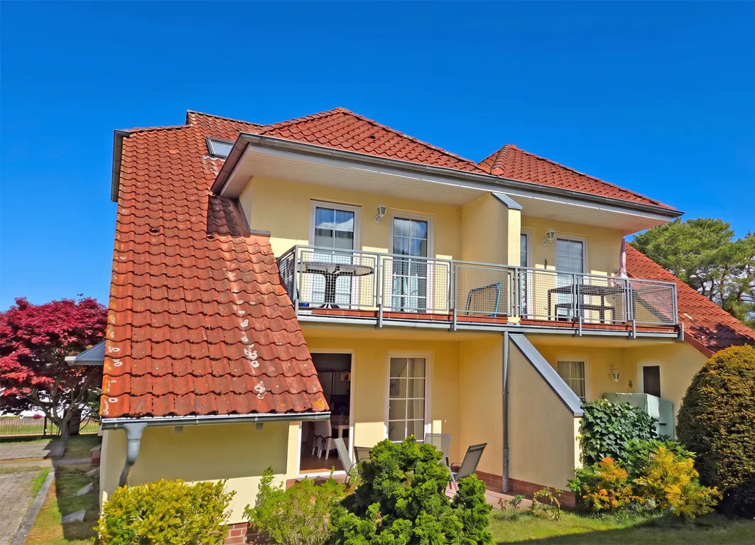 Two-story yellow house with red tile roof, balconies with metal railings, and green landscaping against a clear blue sky.