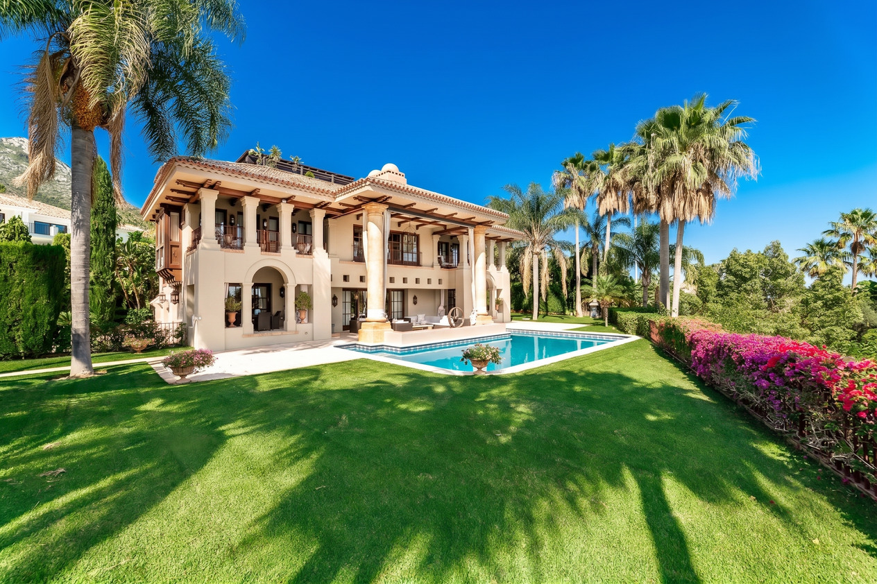 Luxury Mediterranean villa with a pool, lush green lawn, palm trees, and vibrant pink bougainvillea under a clear blue sky.