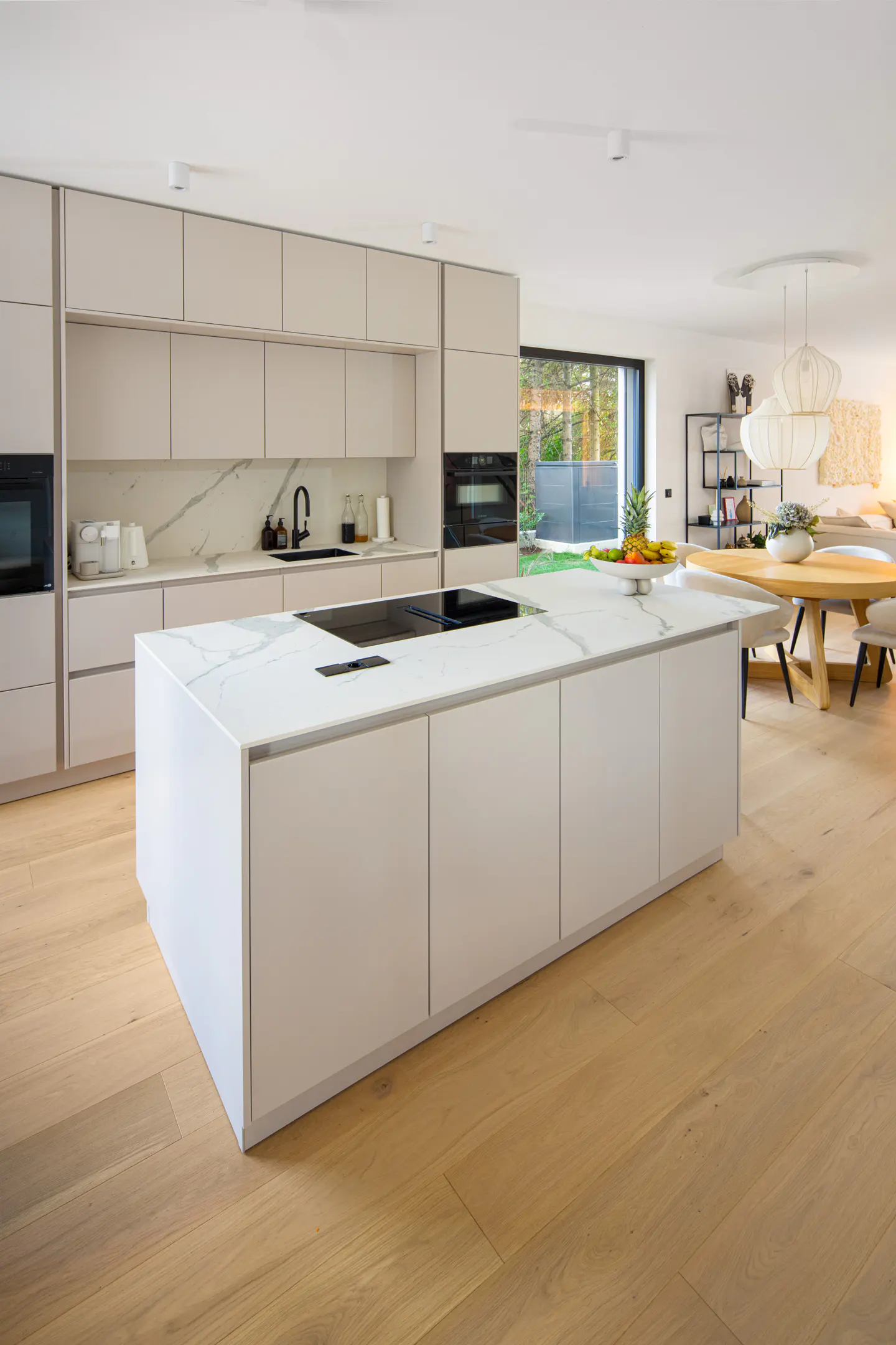 A modern kitchen with light wood floors, white cabinets, and a marble countertop island. A dining area is visible in the background.