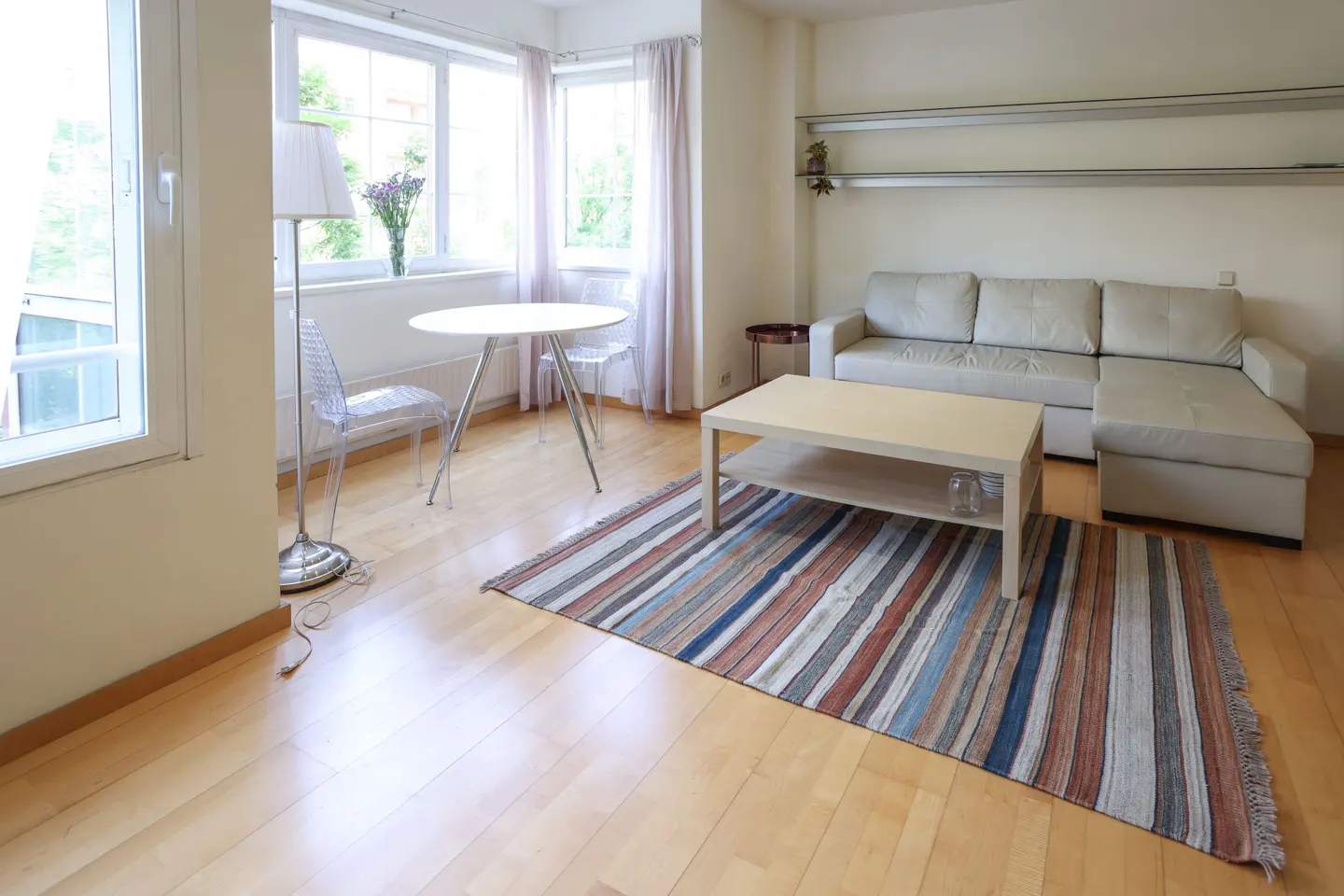 Bright living room with hardwood floors, a striped rug, a beige sofa, and a white table with clear chairs. Large windows let in natural light.