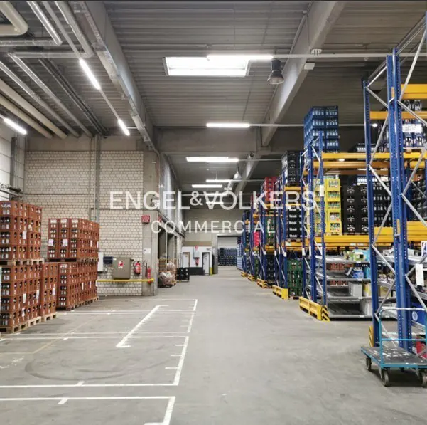 A wide shot of a warehouse interior with tall shelves stocked with goods. Pallets of goods are stacked on the left. The floor is concrete with white lines.