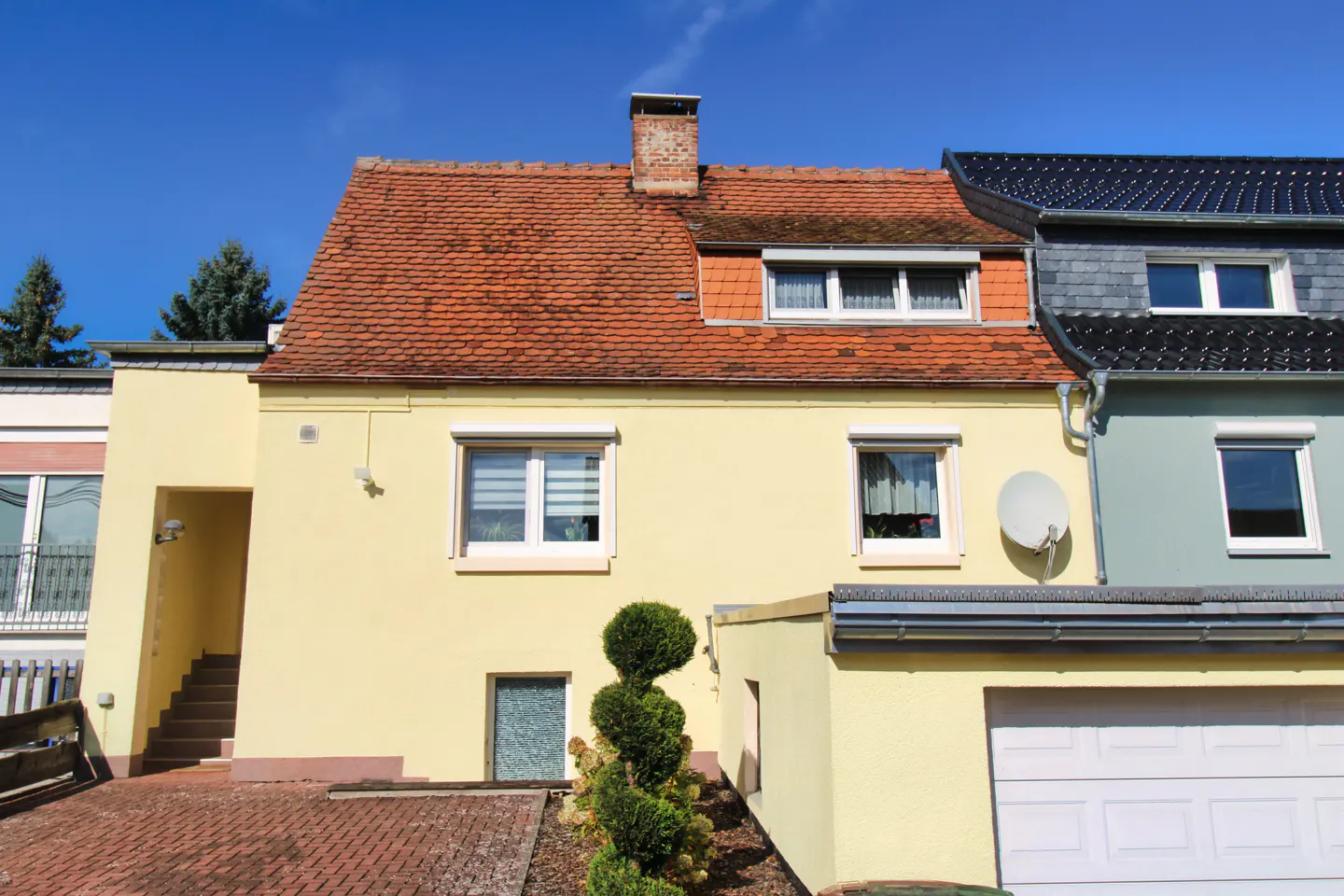 A yellow two-story house with a red tiled roof, a brick chimney, and a white garage door under a clear blue sky.