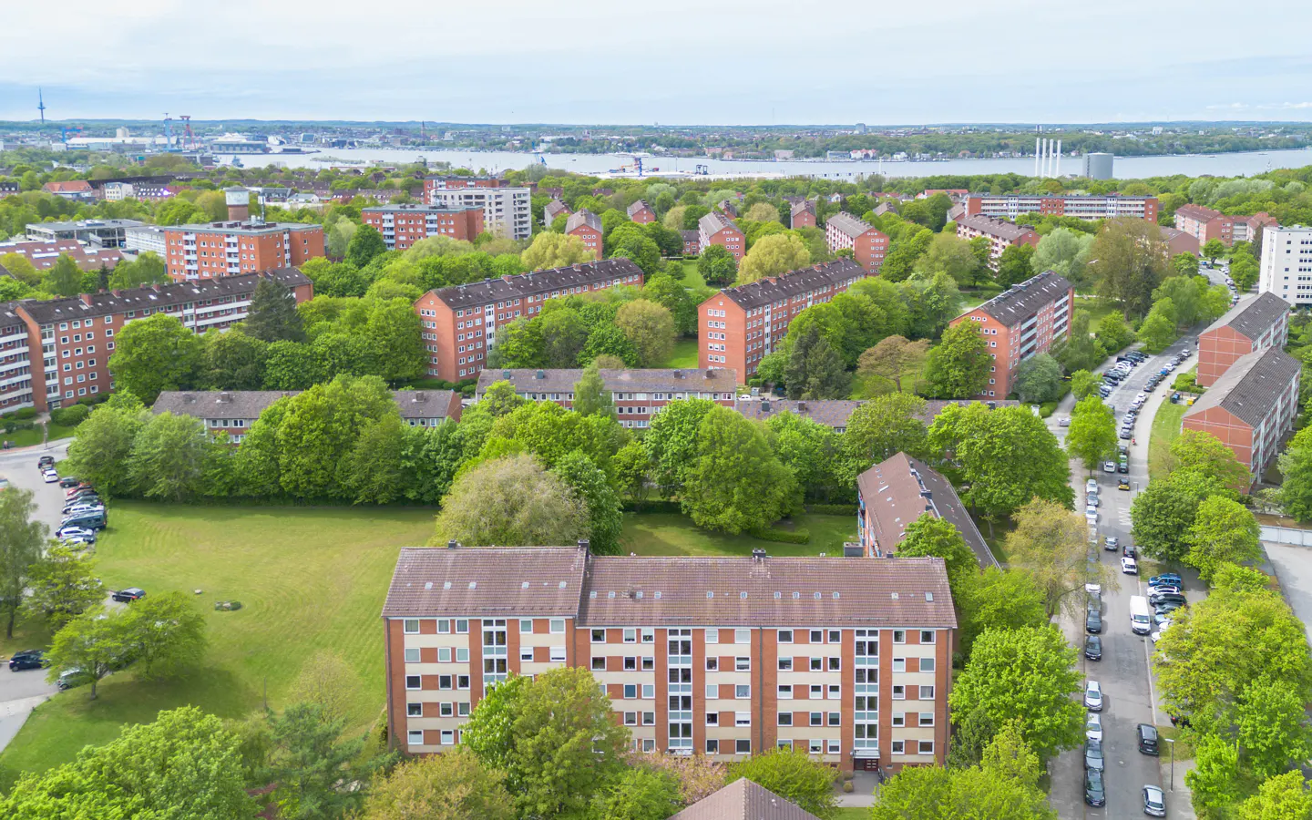 Aerial view of a residential area with red brick apartment buildings surrounded by green trees and a body of water in the background.