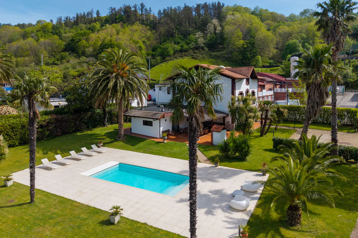 Aerial view of a white house with a red tile roof, a pool, palm trees, and green grass.