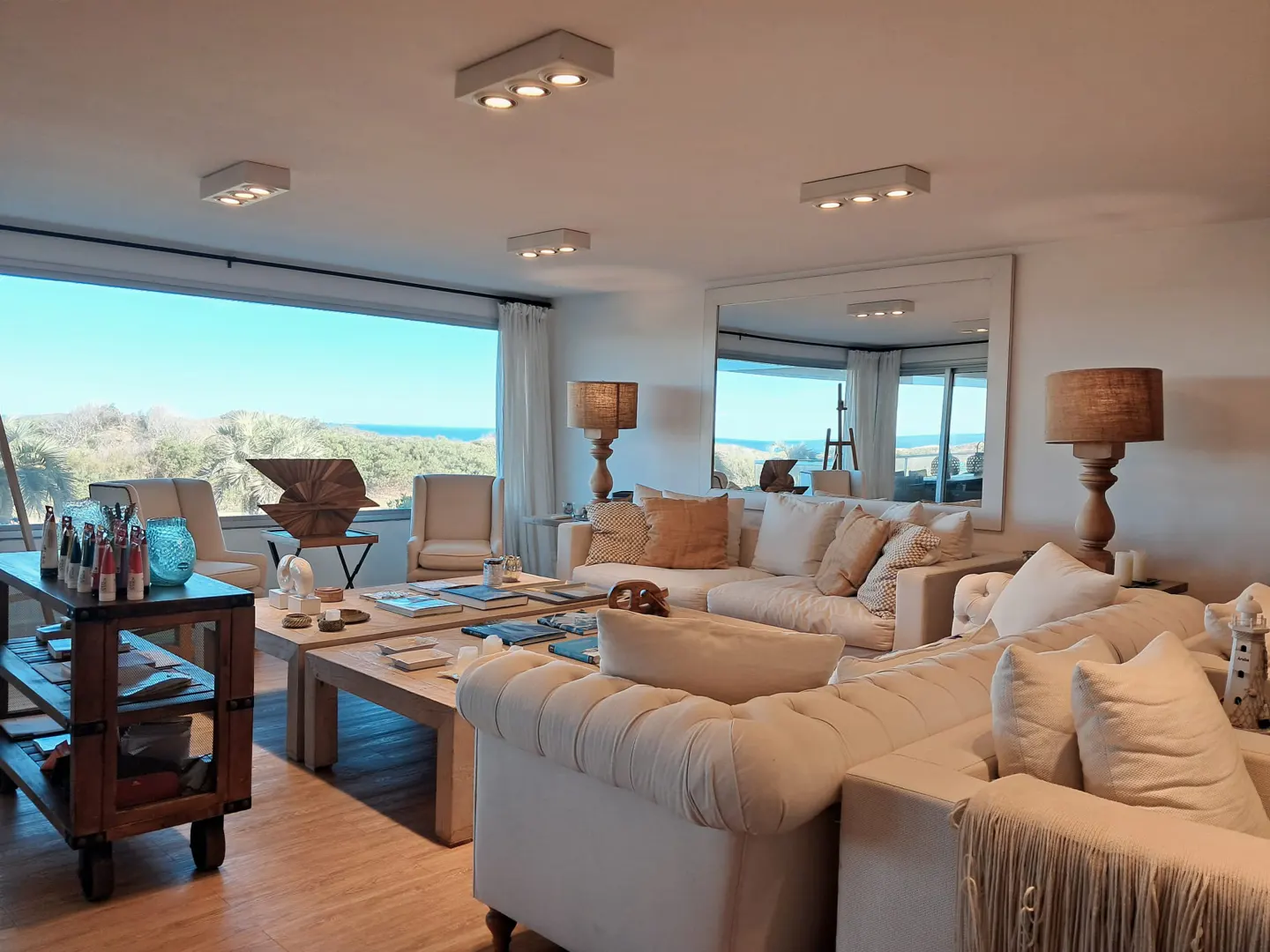 Bright living room with white sofas, wooden tables, and a large window overlooking the ocean. Art supplies and sculptures are visible.