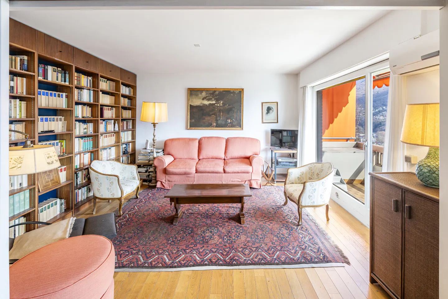 Living room with a large bookcase, pink sofa, two chairs, and a patterned rug on a wood floor. A balcony with an orange awning is visible.