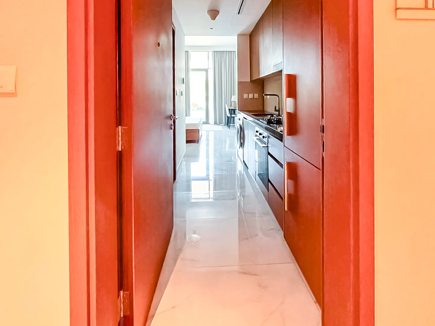View through a doorway into a modern apartment with a sleek kitchen, marble floors, and a bright bedroom in the distance.