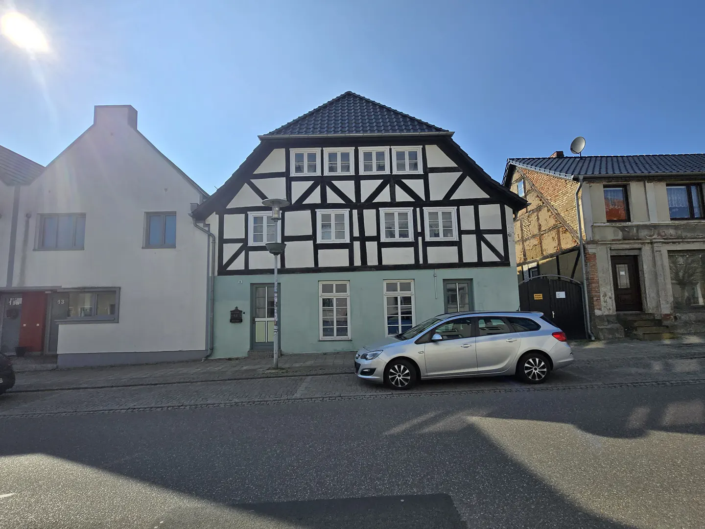 Two-story half-timbered house with a light blue facade, black beams, and white infill, next to a silver car on a sunny day.