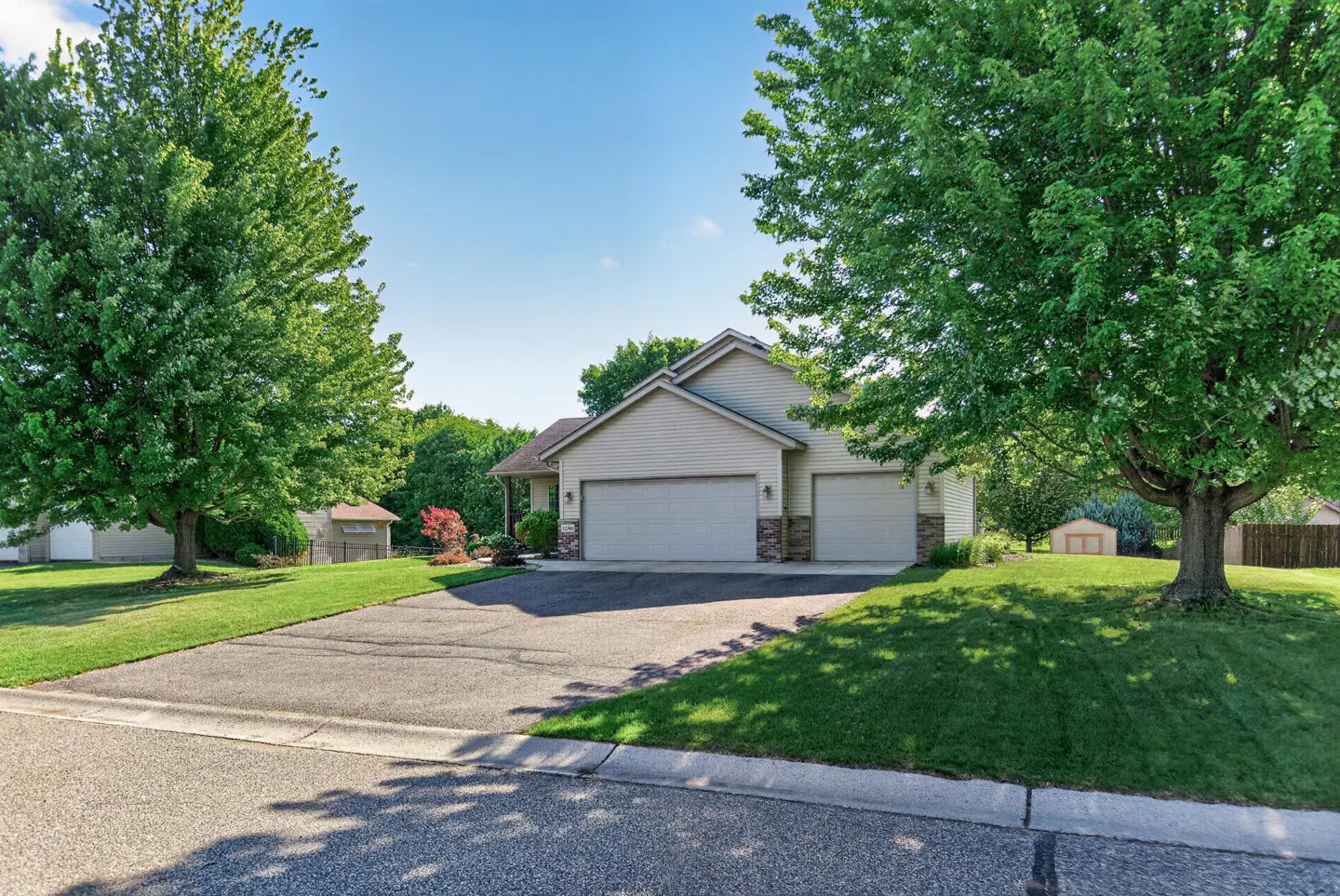 Tan suburban house with a three-car garage, asphalt driveway, and large green trees on a sunny day.