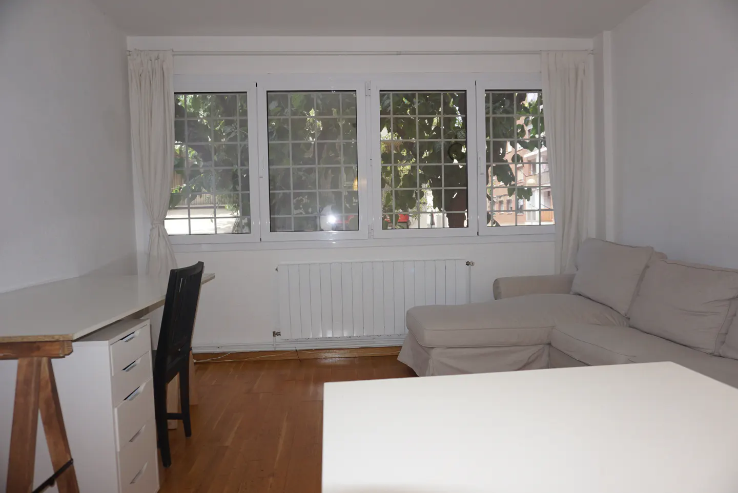 Bright living room with hardwood floors, white walls, and a large window with a view of trees. A desk and sofa are also visible.