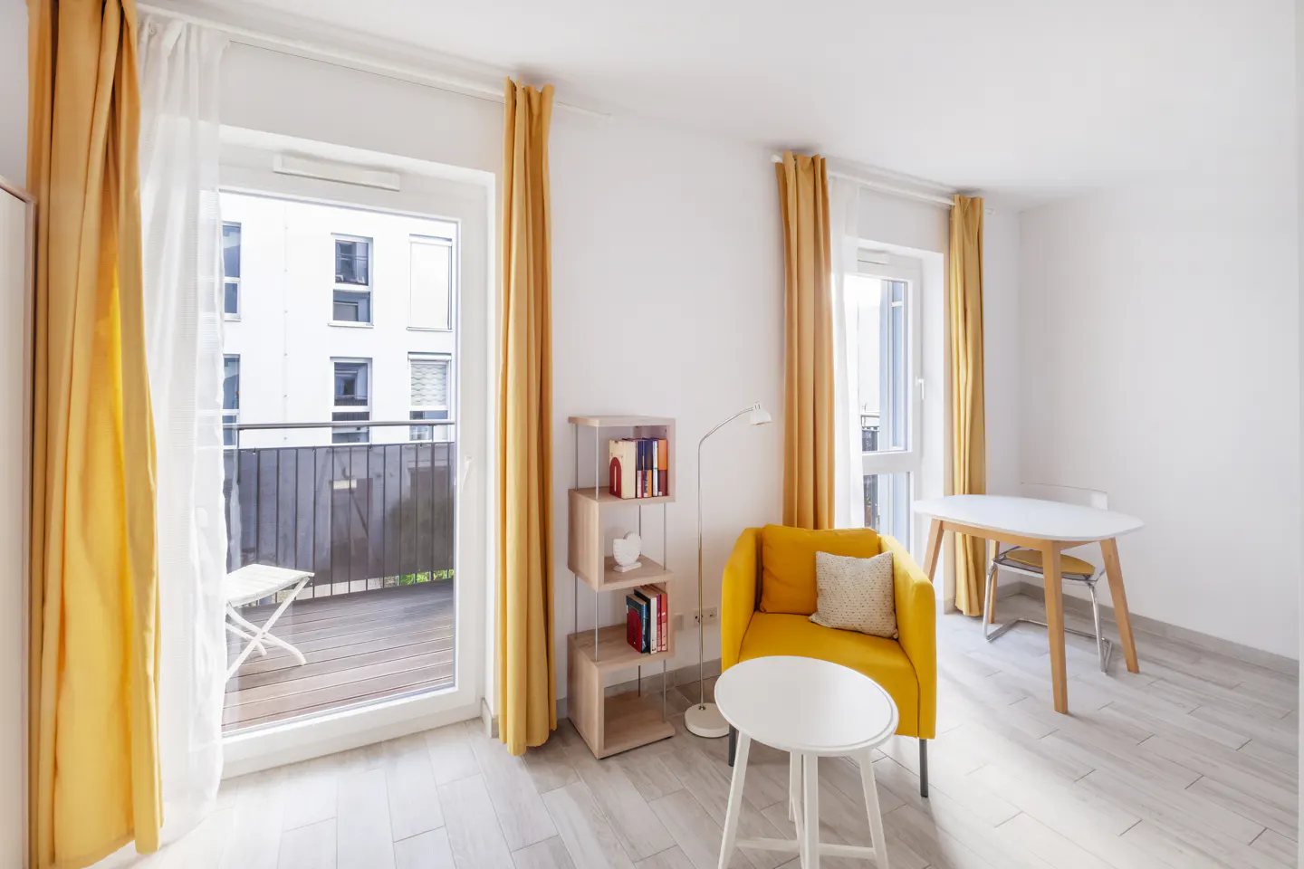 Bright living room with yellow curtains, chair, and white table. Balcony visible through open door. Bookshelf and lamp add detail.