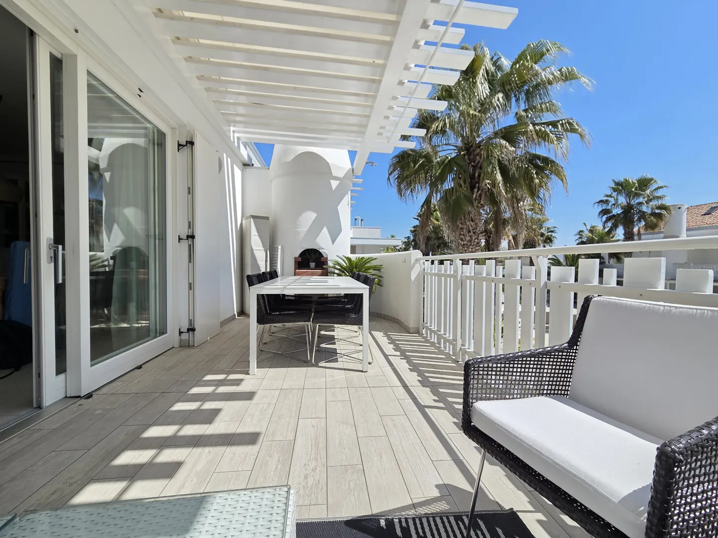 Bright, sunny patio with white pergola, table, chairs, and lounge seating. Palm trees and blue sky visible beyond the white railing.