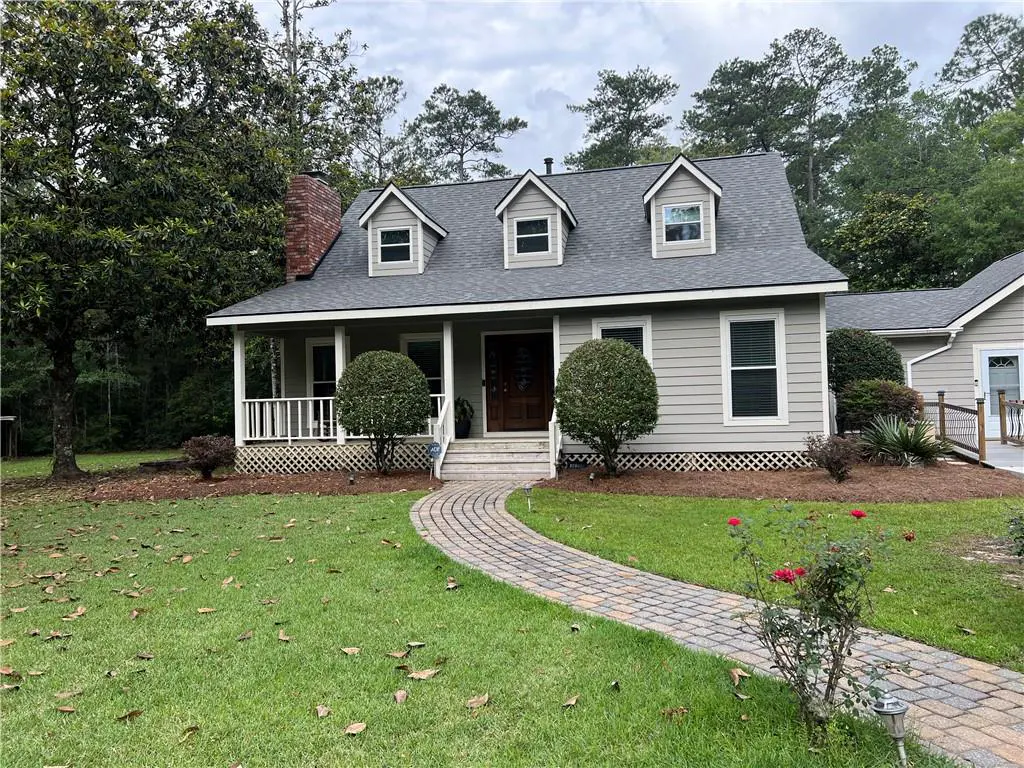 A one-story, light-gray house with a brick chimney and a gray roof. A brick walkway leads to the front door. The lawn is green with trees in the background.
