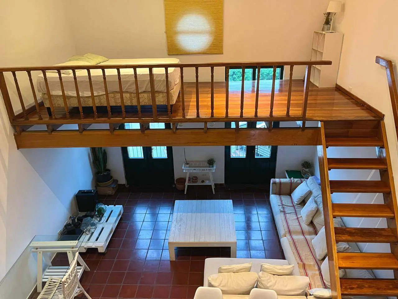 A loft bedroom with a bed overlooks a living room with a white sofa, coffee table, and wooden stairs on tiled floors.