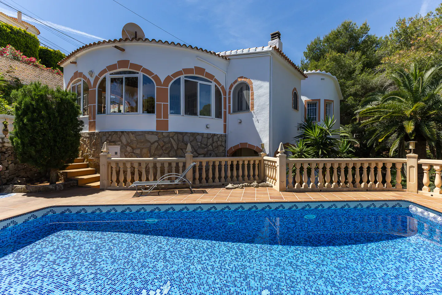 White villa with stone base, arched windows, and a blue tiled swimming pool on a sunny day with palm trees.