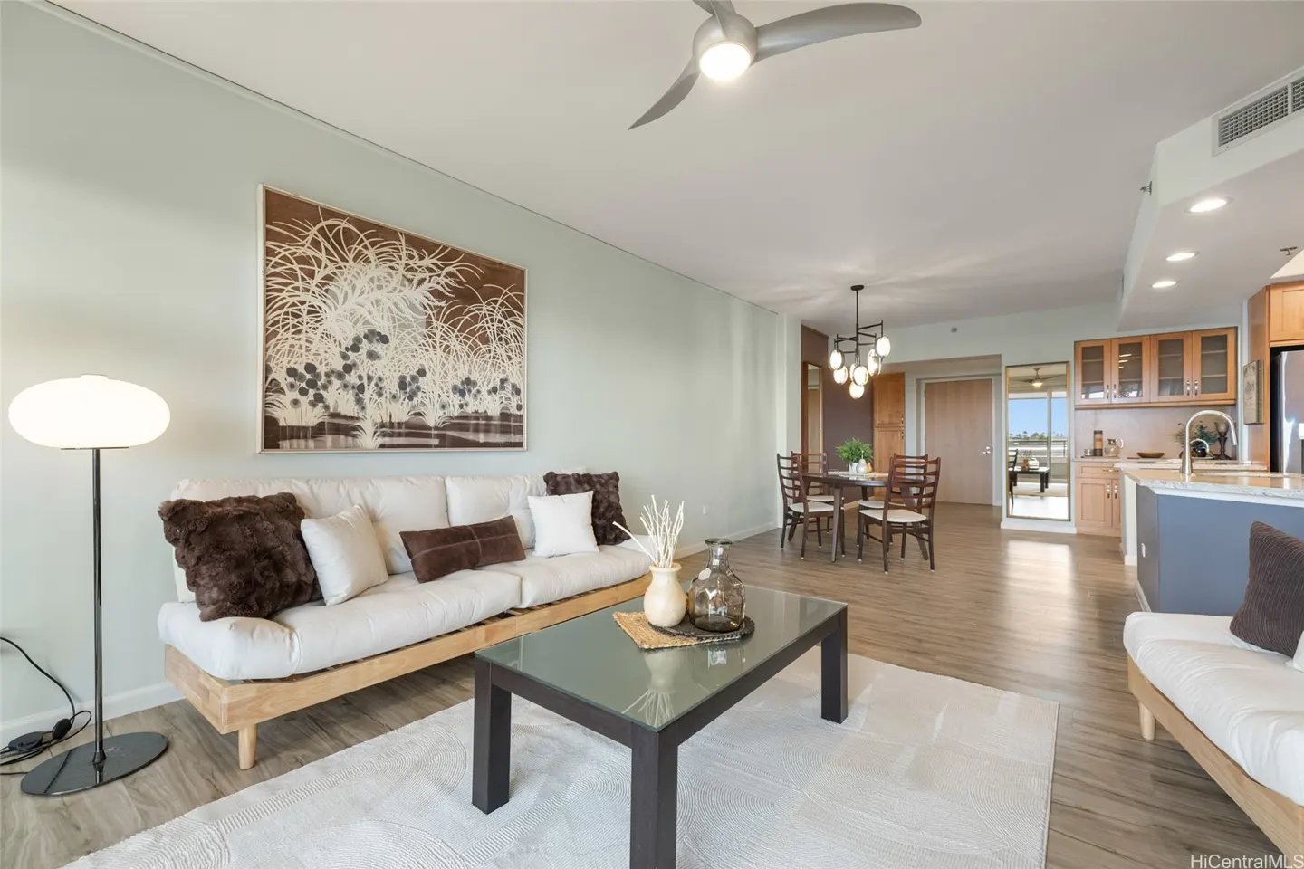 Bright, open-concept living space with a white sofa, glass coffee table, dining set, and kitchen. A ceiling fan is visible.