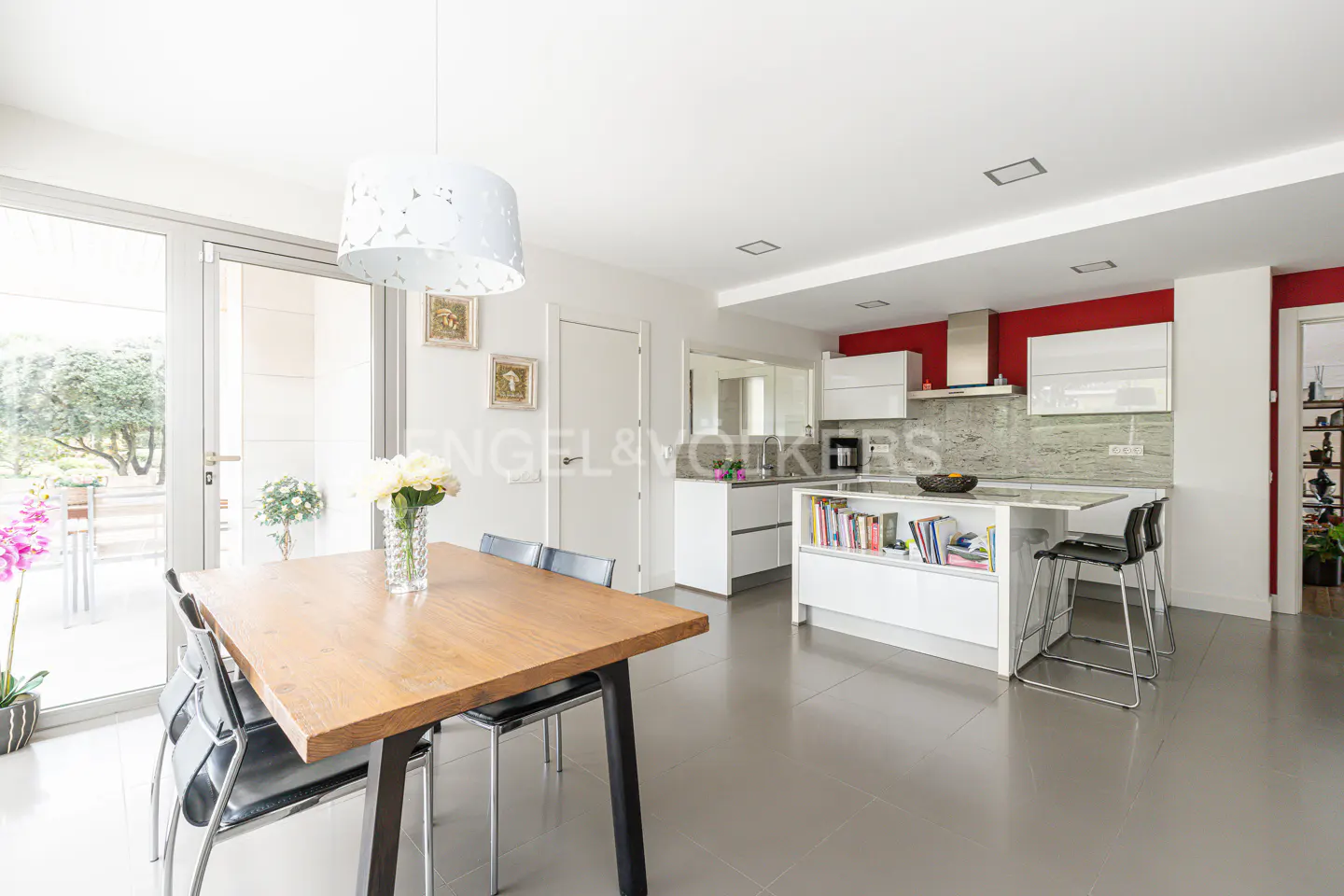 Bright, modern kitchen with a wood table, black chairs, and a white island with seating. Red accent wall behind white cabinets and stainless steel hood.