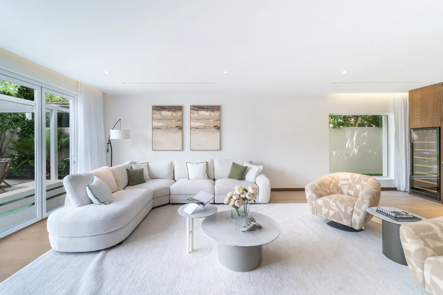 Bright living room with white sectional sofa, patterned armchairs, and round tables on a white rug. Art hangs above the sofa.