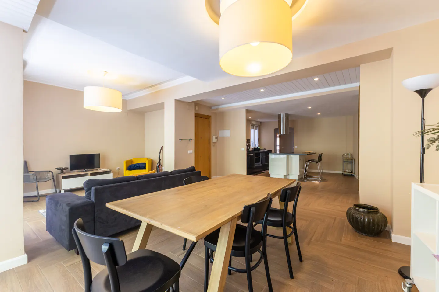 Open-concept living and dining area with a wooden table, black chairs, and a blue sofa. A kitchen is visible in the background.