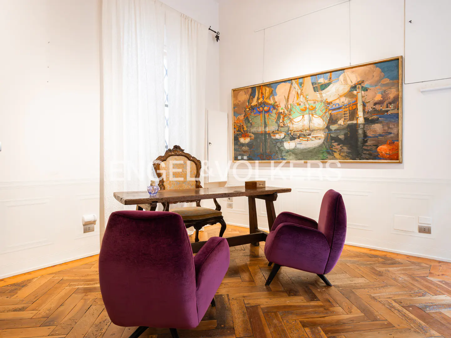 A room with hardwood floors, white walls, and a painting of ships. There is a wooden desk with two purple chairs.
