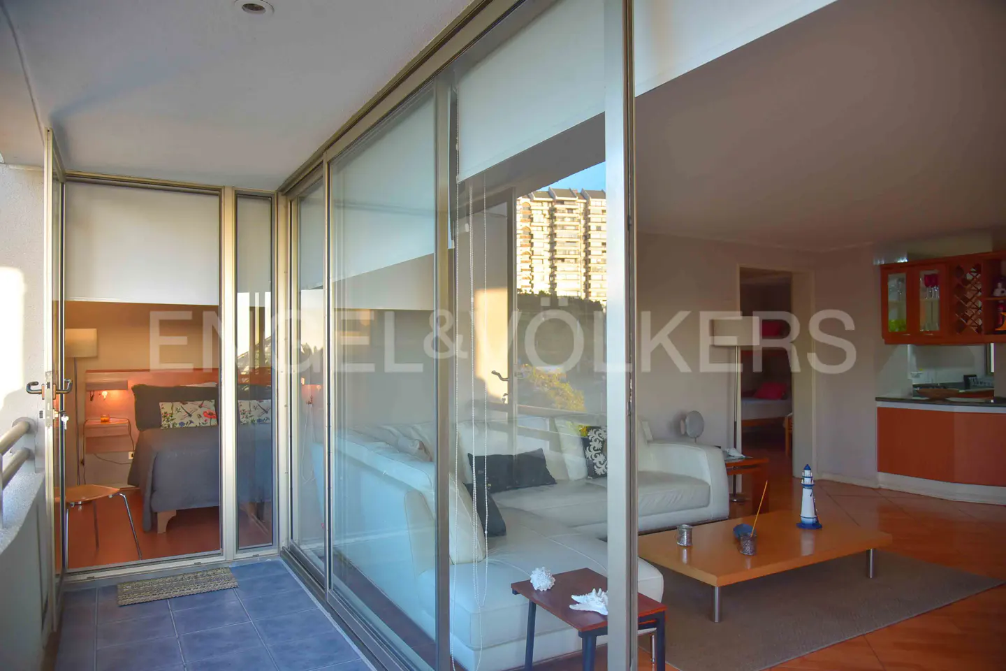 A bright apartment interior with a living room, kitchen, and bedroom visible through glass doors. A white sofa and wooden coffee table are in the living room.