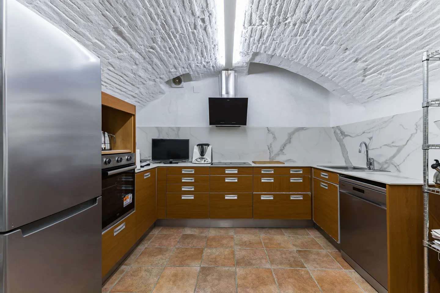 A modern kitchen with brown cabinets, stainless steel appliances, and a white marble backsplash. The floor is tiled in terracotta. The ceiling is arched and made of white brick.