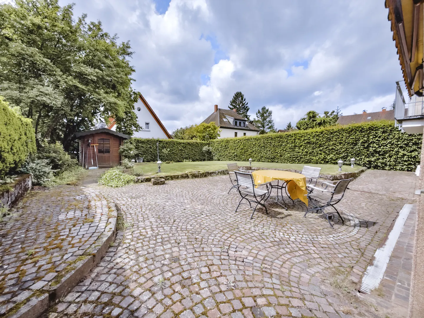 A backyard patio with a stone surface, a table with a yellow cloth, and chairs. A green hedge and trees surround the yard.