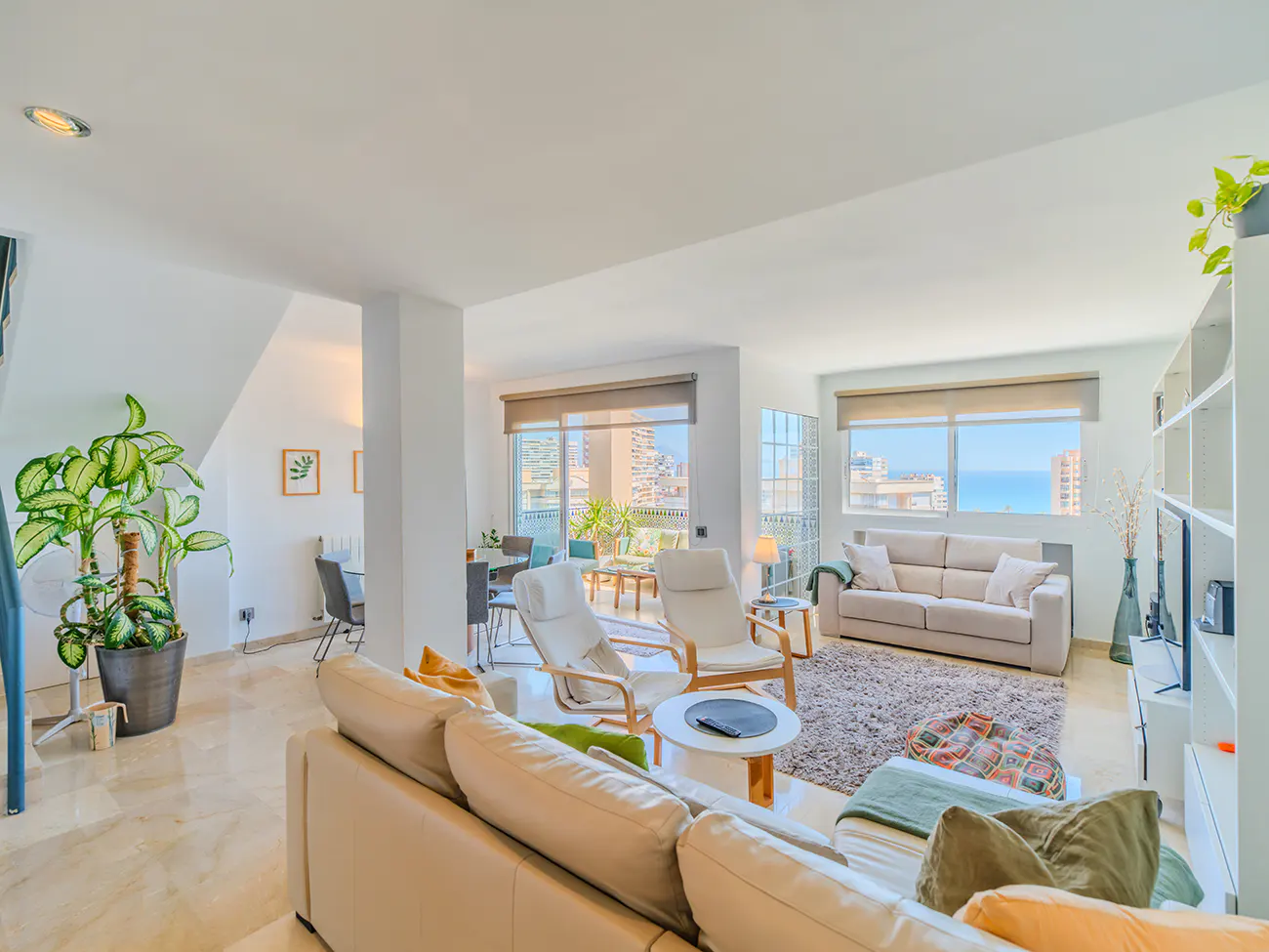 Bright, open-plan living room with beige sofas, white walls, and marble floors. Large windows offer a city and ocean view. Plants add a touch of nature.