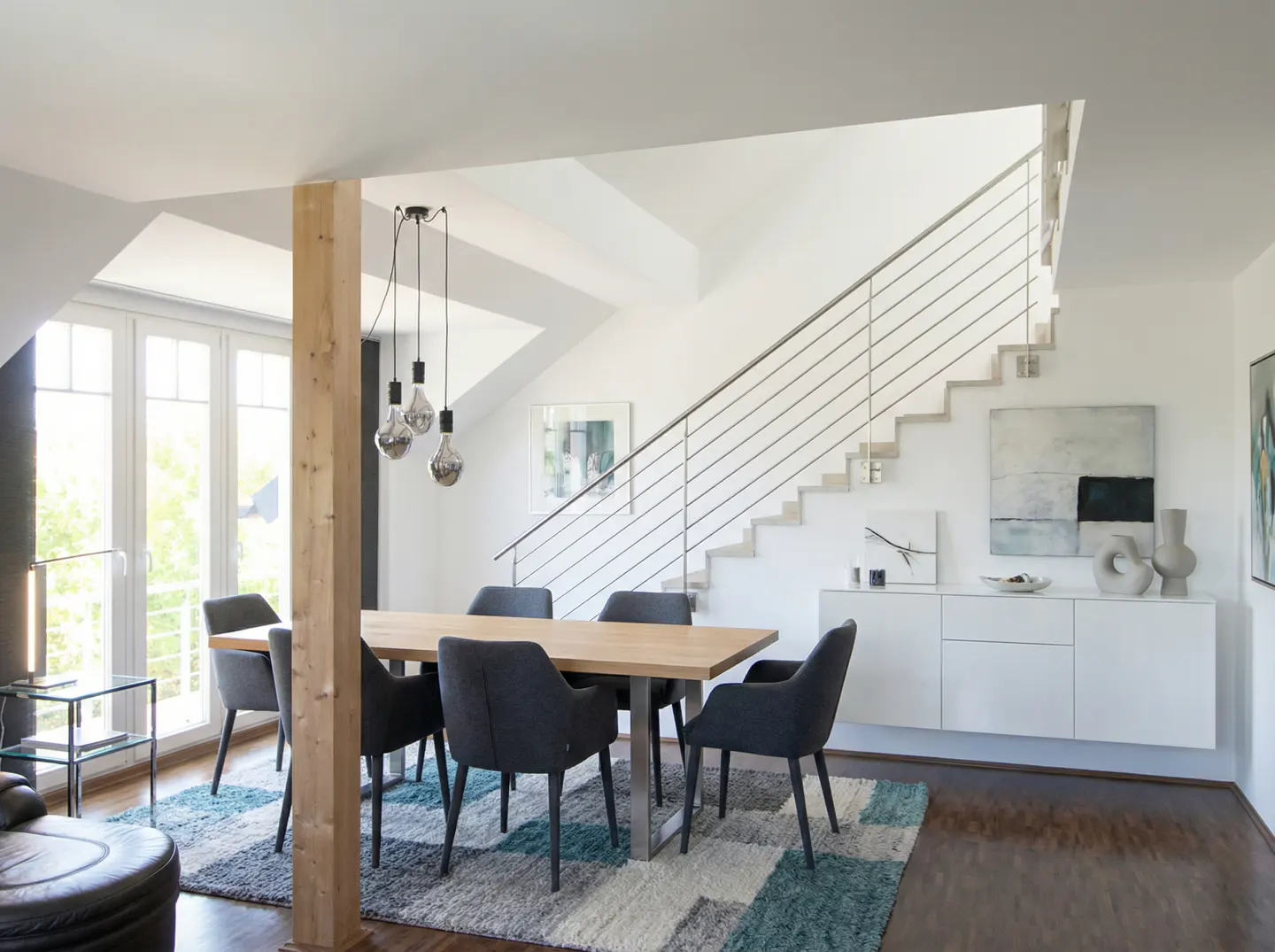 Bright, modern dining room with a wooden table, gray chairs, and a patterned rug. A staircase with metal railings is visible in the background.
