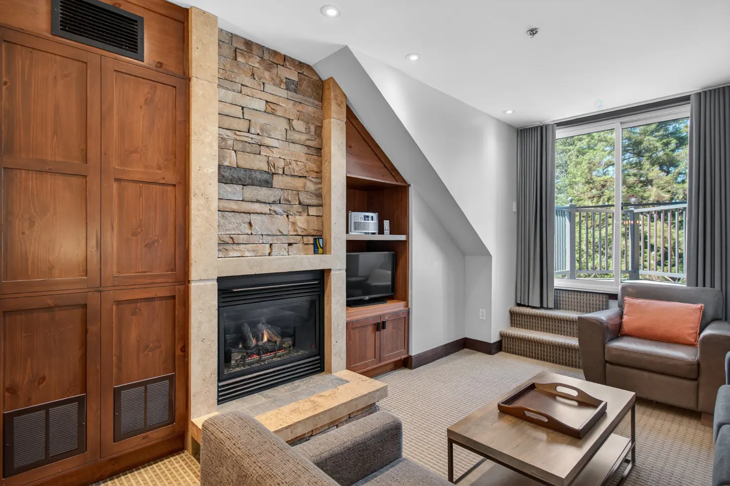 Living room with stone fireplace, wood cabinets, and a window view of trees. A coffee table and armchairs sit on a beige carpet.