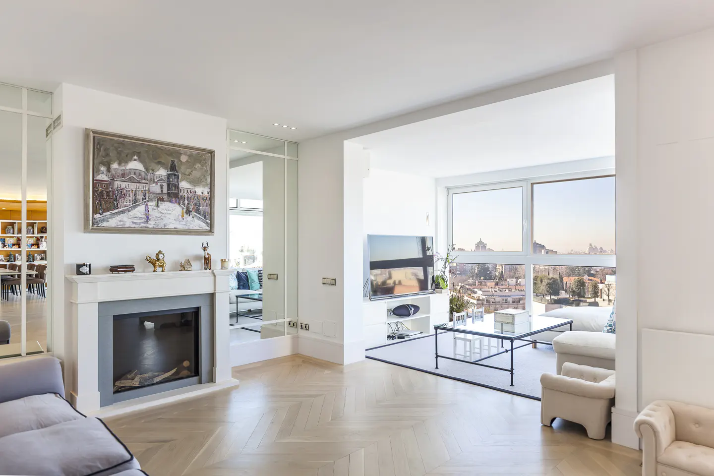 Bright, modern living room with herringbone wood floors, a fireplace with a painting above, and a large window with a city view.