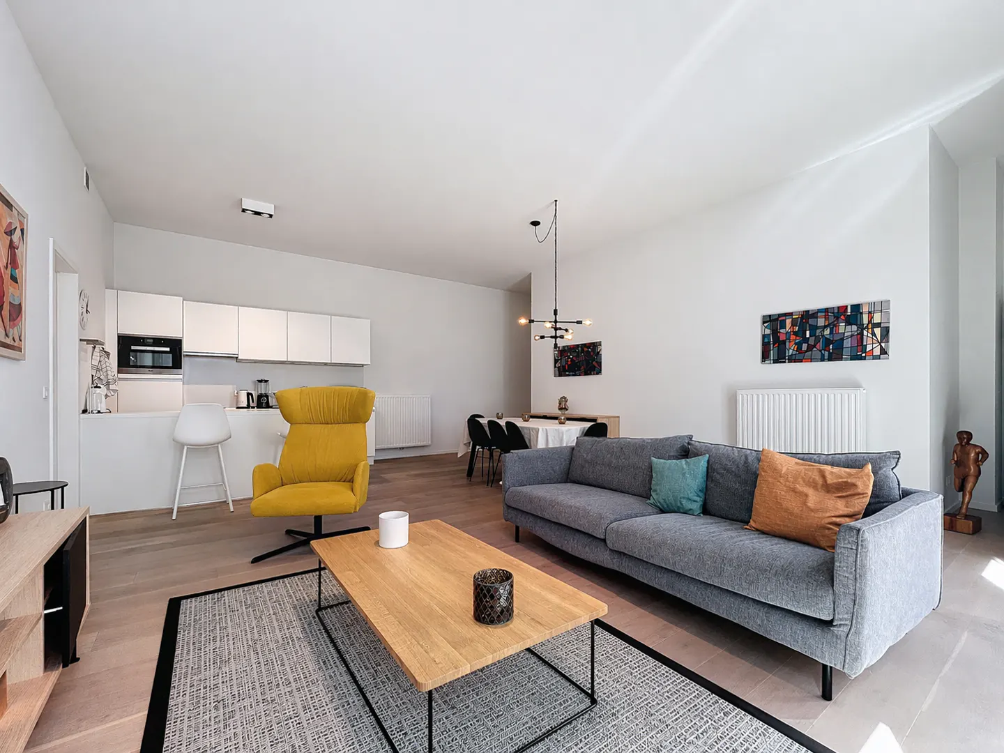 Bright, modern living space with a gray sofa, yellow chair, and wood coffee table on a gray rug. White kitchen in the background.