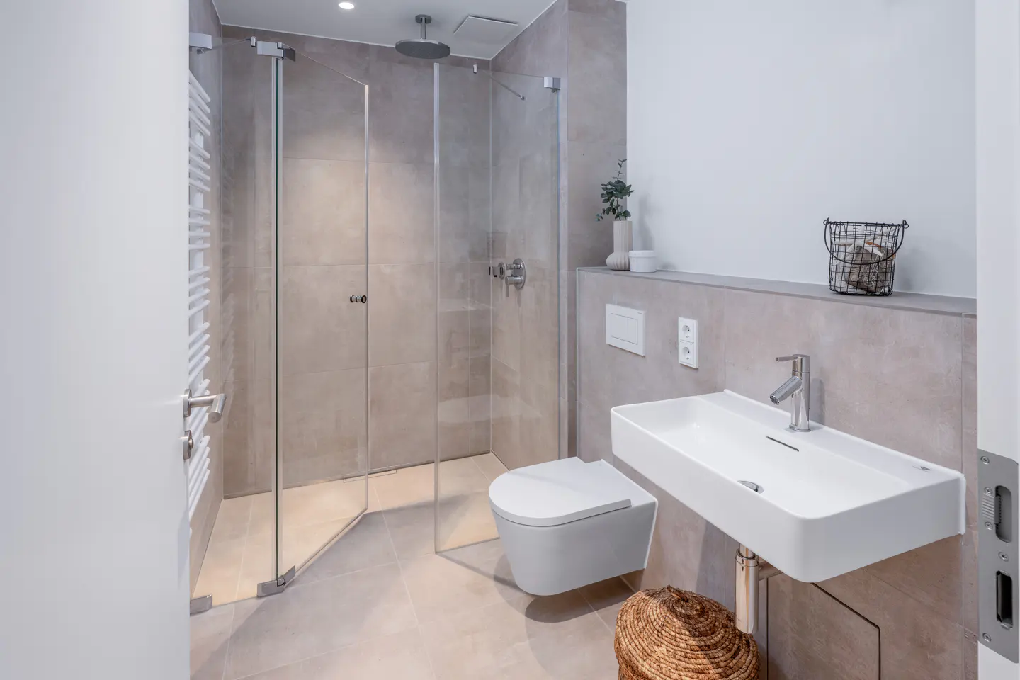Modern bathroom with a glass shower, white toilet and sink, and beige tile. A towel warmer is on the left.
