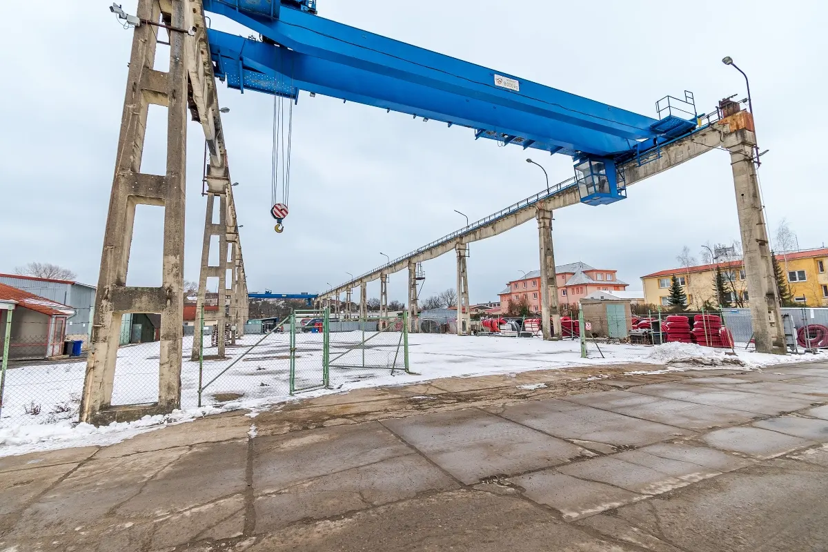 Outdoor shot of a large blue gantry crane with a hook, supported by concrete pillars in a snowy industrial yard.