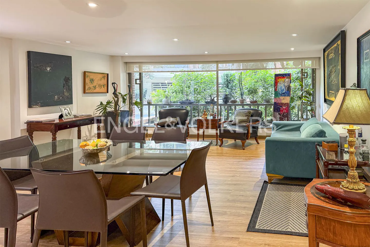 Bright living room with a glass table, brown chairs, a blue sofa, and a large window overlooking a green garden. Art adorns the walls.