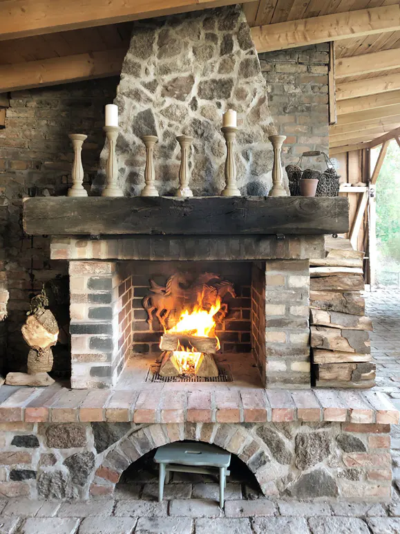 Stone fireplace with burning logs, topped with a wooden beam and six white candle holders. A small blue stool sits in the arched base.