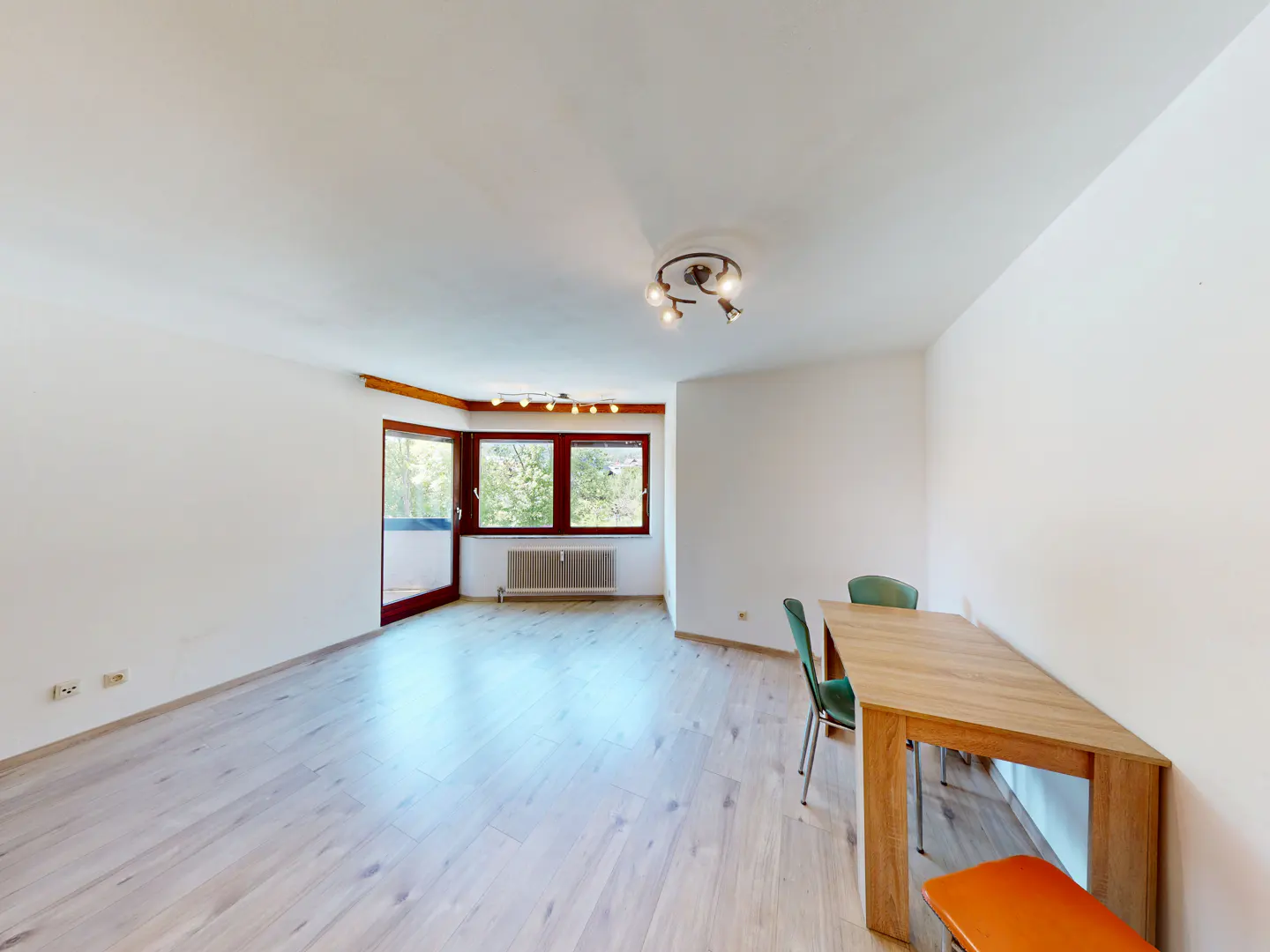 Bright, empty room with light wood floors, white walls, and a wood table with green chairs. Windows and a door lead to a balcony.