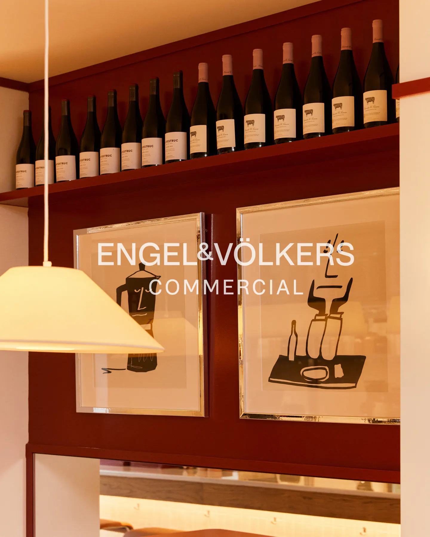 Interior view of a commercial space with wine bottles on a shelf, framed art, and a hanging lamp.