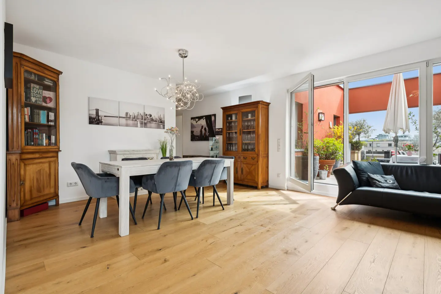 Bright, modern living room with wood floors, white walls, and a white table with blue chairs. A balcony with a black sofa is visible through open doors.