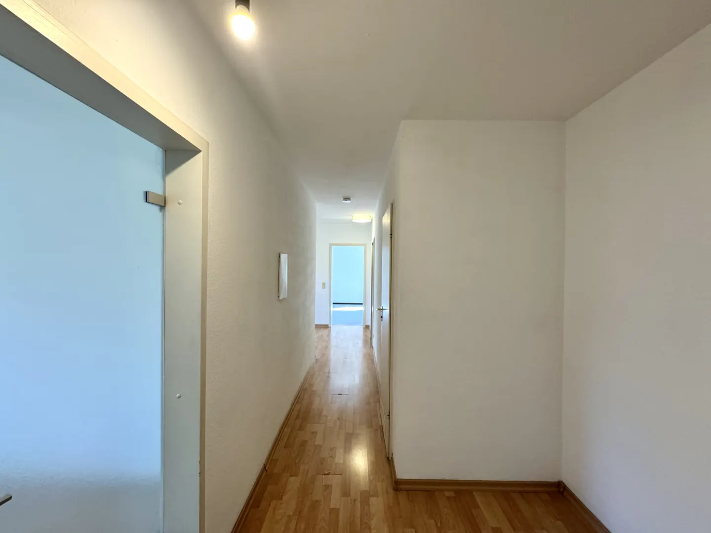 A bright hallway with wood floors and white walls leads to a blue room in the distance.