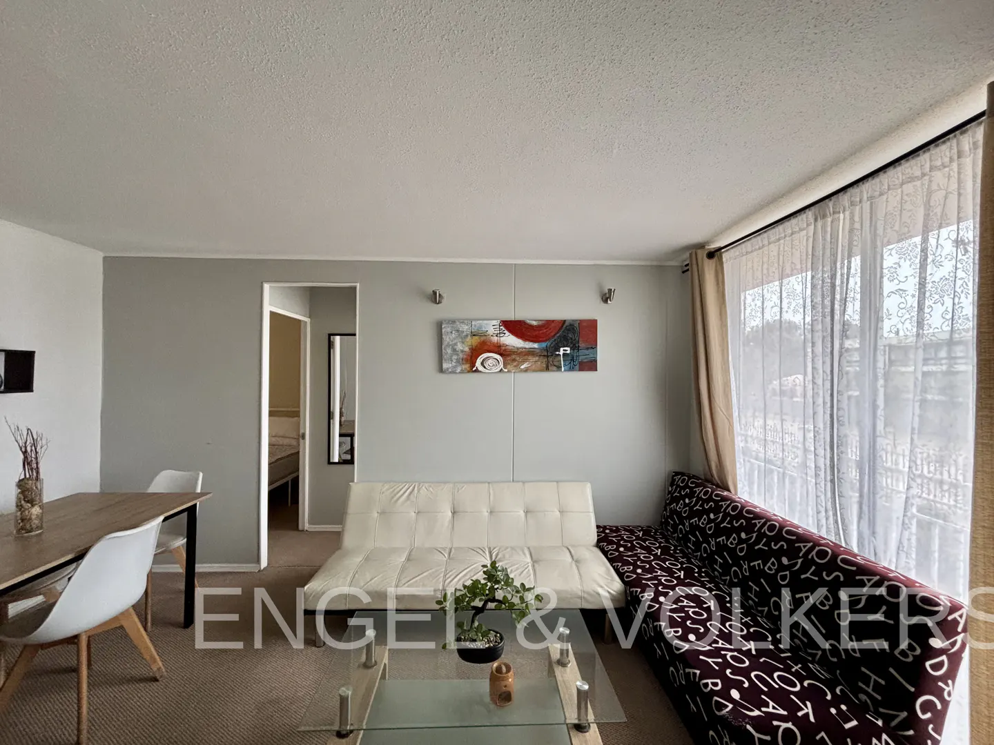Living room with a dining table, white futon, patterned sofa, and glass coffee table. Gray walls and sheer curtains. Engel & Volkers logo.