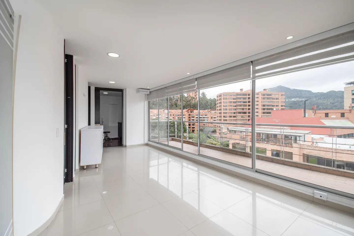 Bright, empty room with white walls and tile floor. Large windows offer a city view with buildings and mountains in the distance.